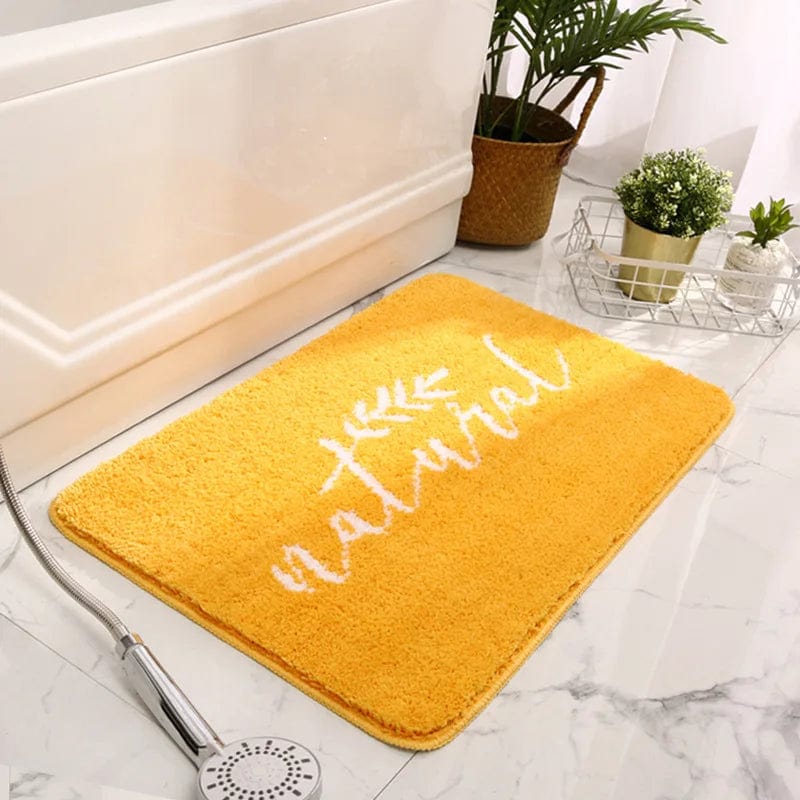 Yellow natural bath mat with white script text, placed beside bathtub on marble floor.
